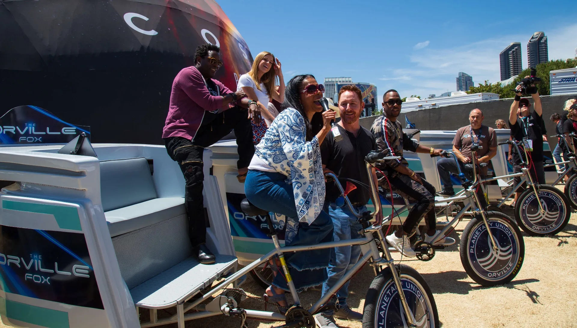 Orville cast riding pedicab fleet past COSMOS dome with San Diego skyline
